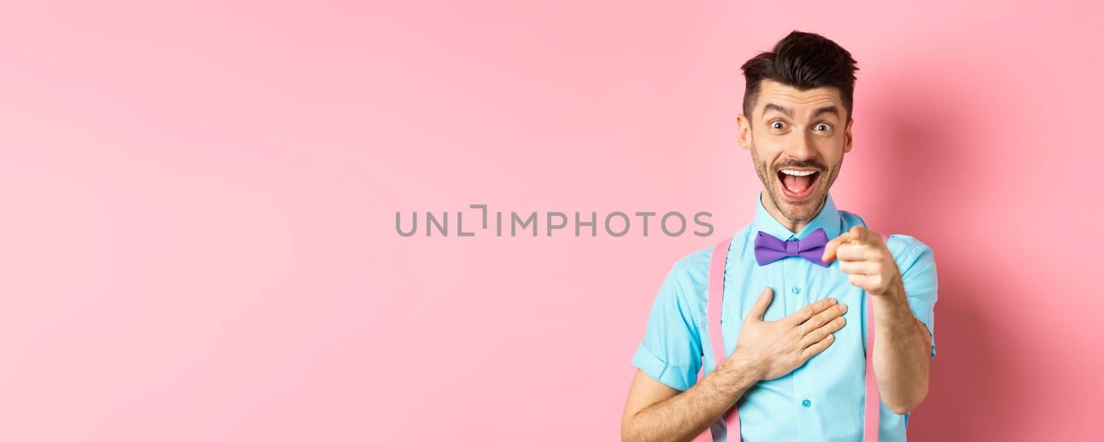 Funny guy laughing at something hilarious, smiling amazed and pointing finger at camera, standing on pink background.