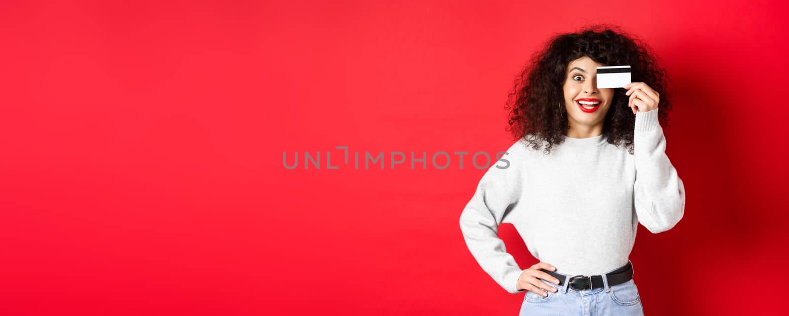 Stylish girl with curly hair showing plastic credit card over eye and smiling, standing against red background.