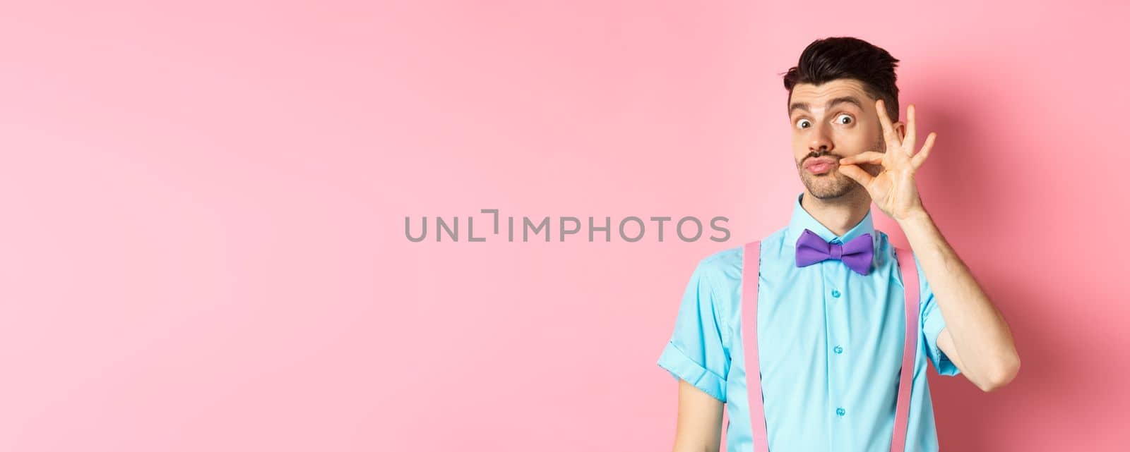 Funny young man touching his french moustache and pucker lips, looking silly at camera, standing in bow-tie and suspenders on pink background.