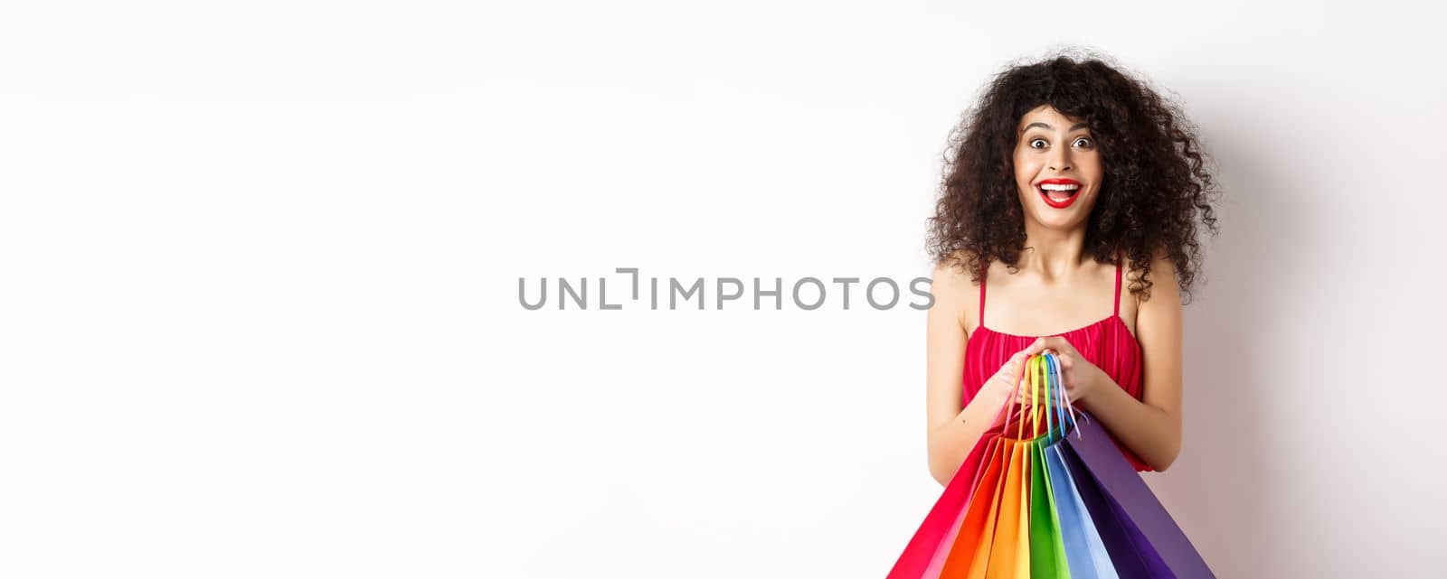 Cheerful young woman in stylish red dress, holding shopping bags and smiling, buying during promo offers, standing over white background.