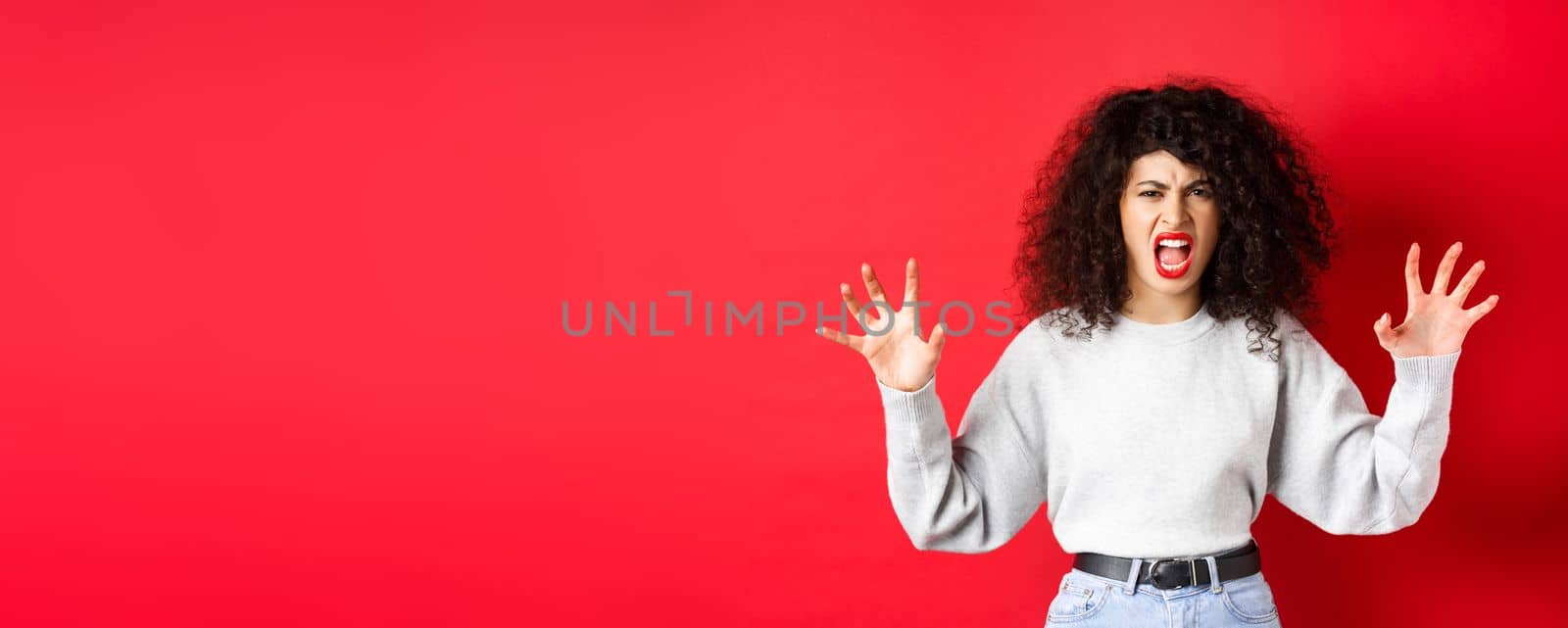 Scary woman trying to scare you, shouting and showing animal claws gesture, screaming at person, standing against red background.