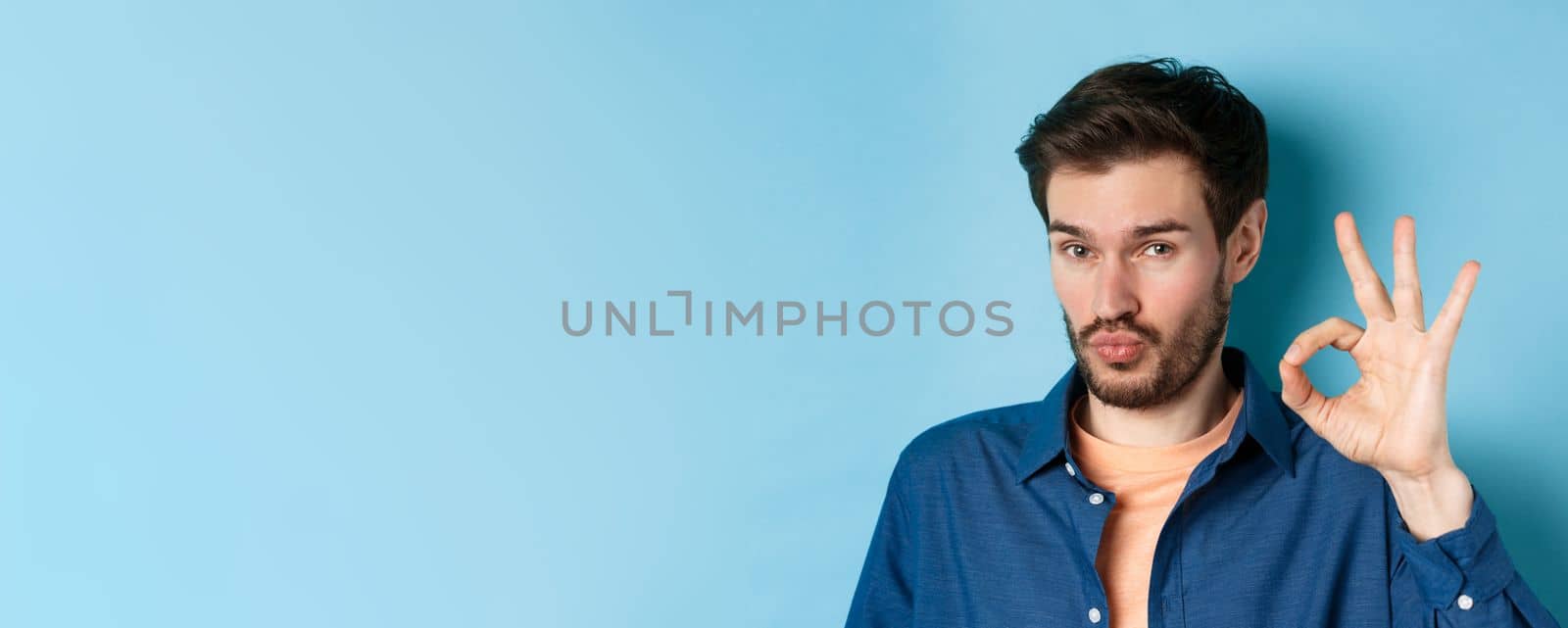 Image of silly young man pucker lips and show okay gesture, praise good thing, compliment excellent choice, standing on blue background.