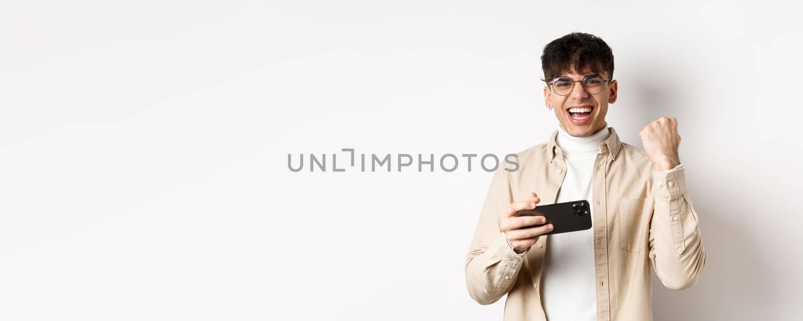 Happy guy winning on smartphone, holding mobile phone and raising hand up, shouting yes with joy, standing on white background.