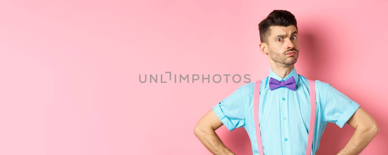 Image of young man with funny moustache and bow-tie standing in confident pose, staring with disbelief and doubts at camera, posing over pink background.