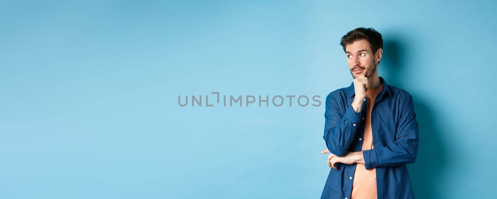 Thoughtful caucasian man looking aside at empty space and thinking, standing pensive on blue background. Copy space