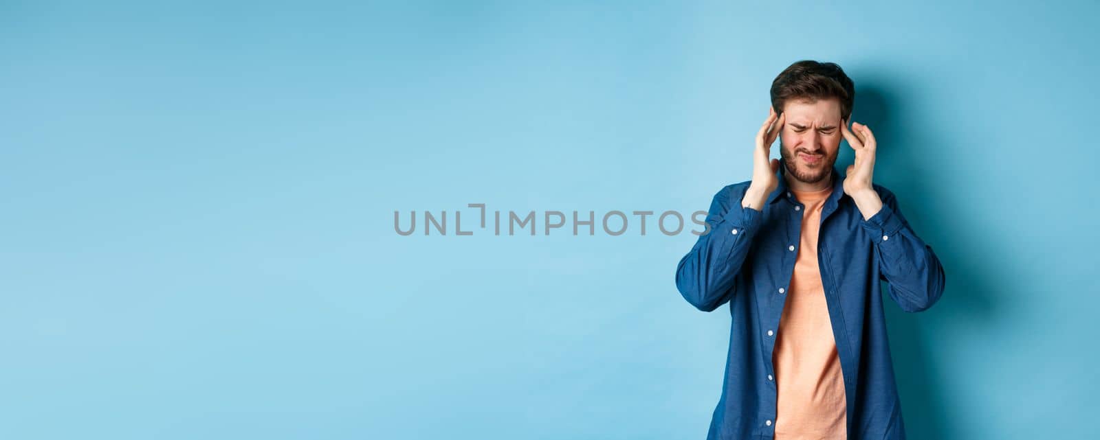 Man feeling sick, touching head and grimacing from painful migraine, having headache or feel dizzy, standing on blue background.