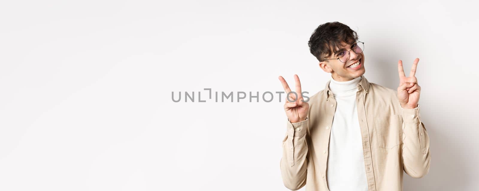 Real people. Handsome hipster guy in glasses showing peace gestures and smiling cute, standing on white background.