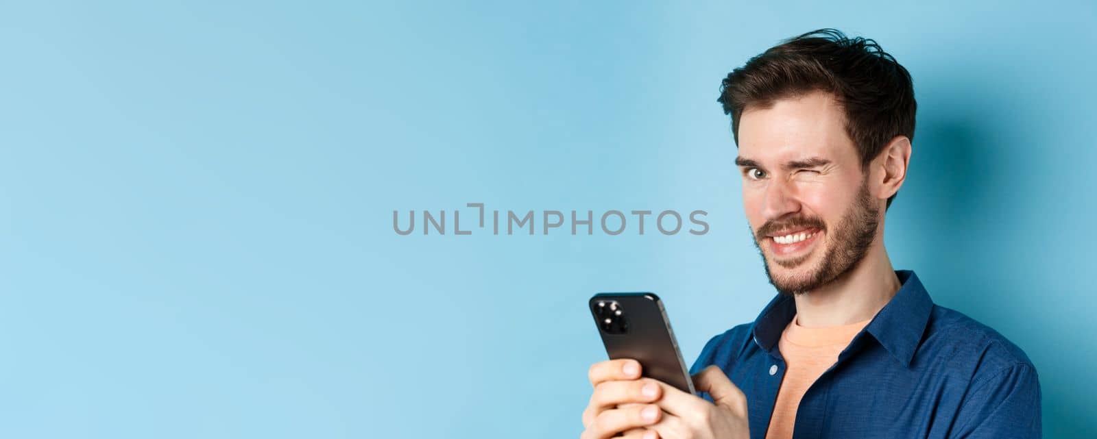 Portrait of handsome young man with beard, winking and smiling at camera, using smartphone, standing on blue background by Benzoix