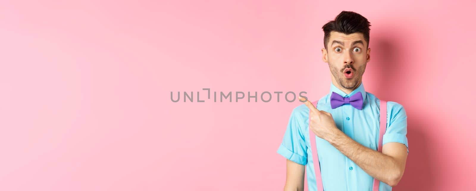 Check it out. Amazed caucasian man with moustache, wearing bow-tie, saying wow and pointing left, showing advertisement, standing on pink background by Benzoix