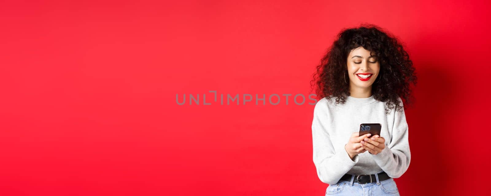 Happy positive girl chatting on phone, reading message and smiling, using social media app, standing on red background by Benzoix