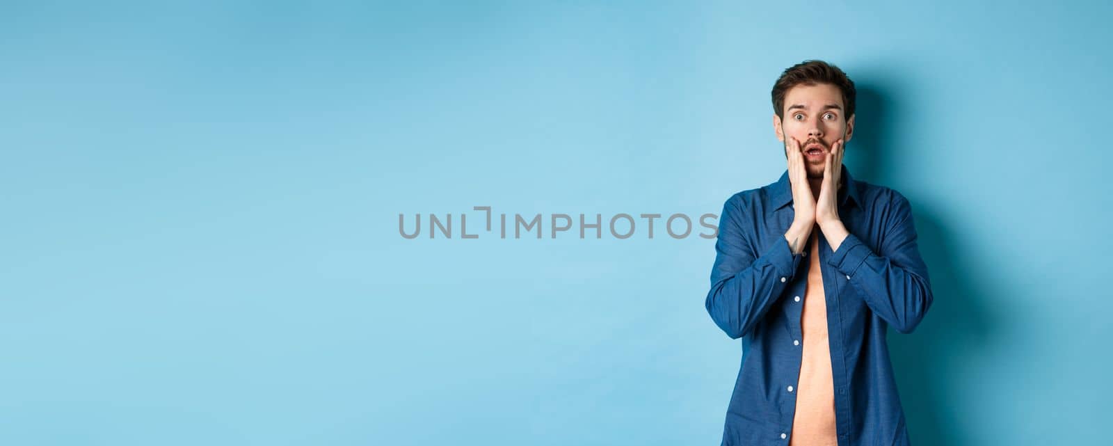 Portrait of guy gasping and dropping jaw with shocked face, staring at camera startled, standing on blue background by Benzoix