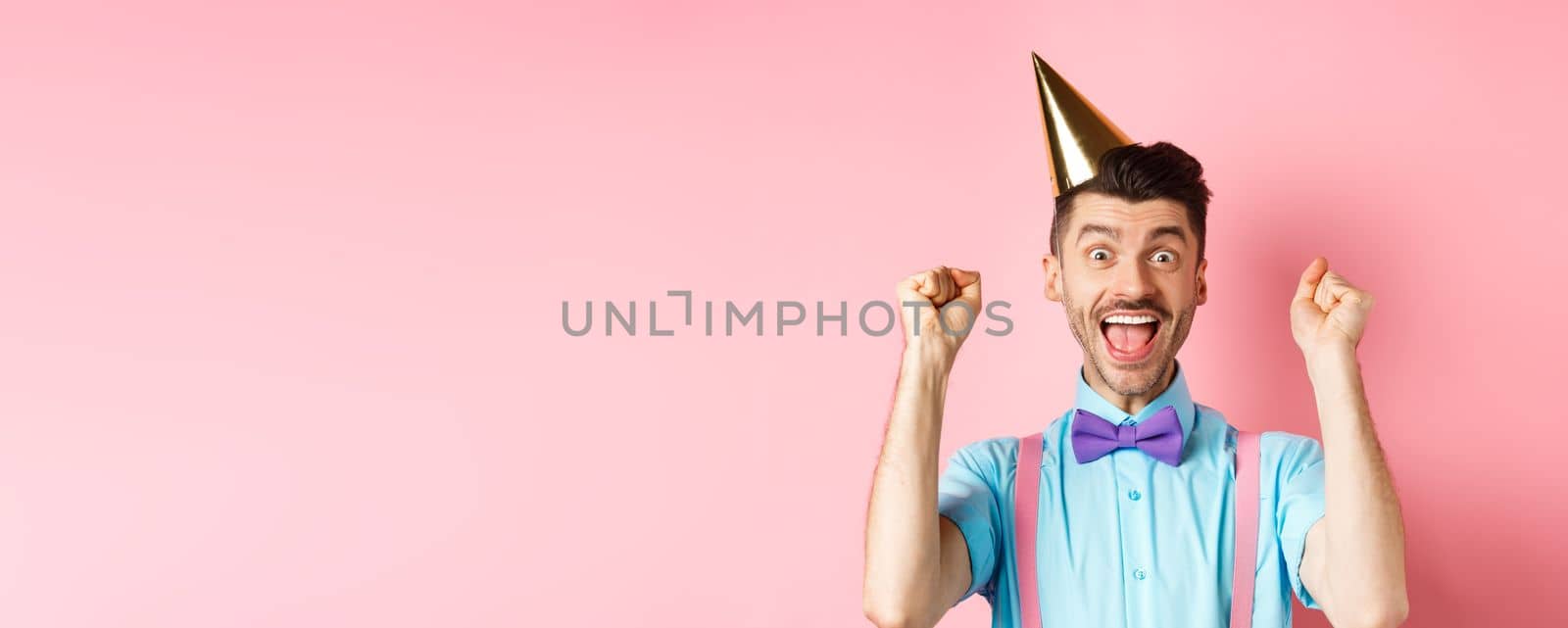 Holidays and celebration concept. Cheerful young man celebrating birthday party in cone hat and bow-tie, shouting yes with excitement and making fist pump gesture, pink background by Benzoix