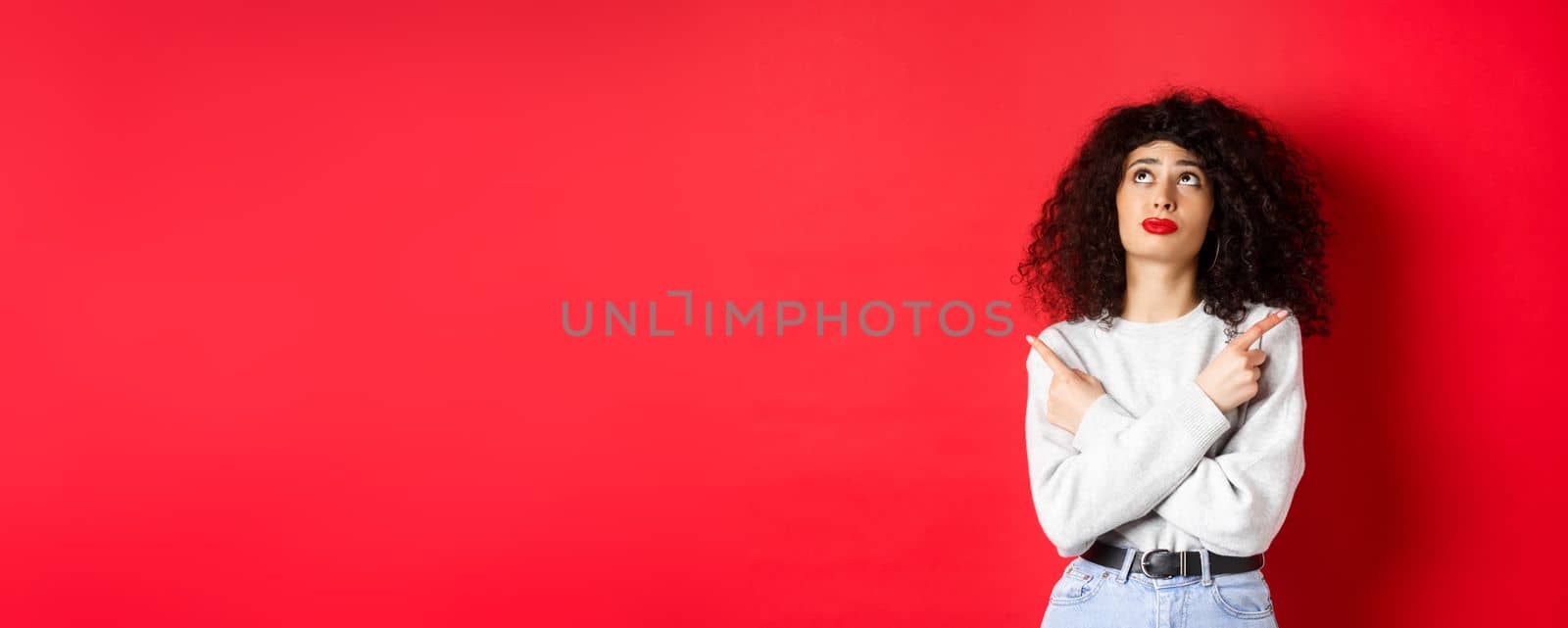 Indecisive female model with red lips and curly hair, looking up troubled with making choice, pointing fingers sideways, choosing between two products, studio background.