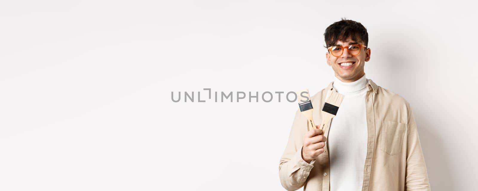 Hobbies and leisure concept. Stylish young man in glasses showing painting brushes and smiling, inviting to paint something, standing on white background.