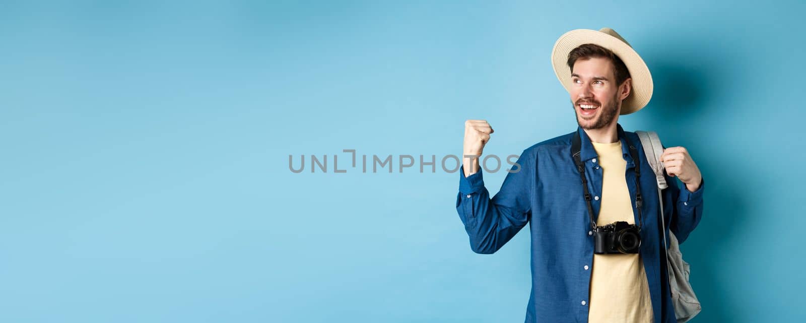 Cheerful tourist rejoicing on vacation, celebrating summer holidays, showing fist pump gesture and saying yes with satisfied face, looking aside, blue background.