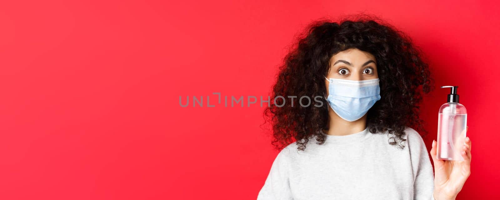 Covid-19, pandemic and quarantine concept. Excited girl with curly hair, wearing medical mask, showing bottle of hand sanitizer or antiseptic, standing on red background.
