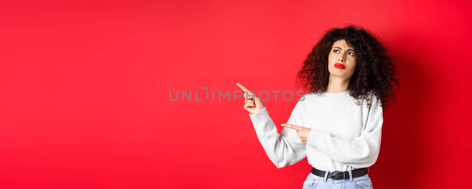 Skeptical frowning girl with curly hair, pointing and looking left hesitant, standing upset on red background.