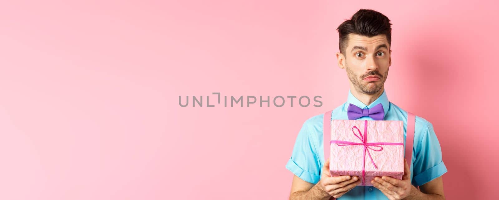 Holidays and celebration concept. Attractive young man with moustache, wearing festive outfit with bow-tie, showing cute gift box and looking at camera, standing over pink background.