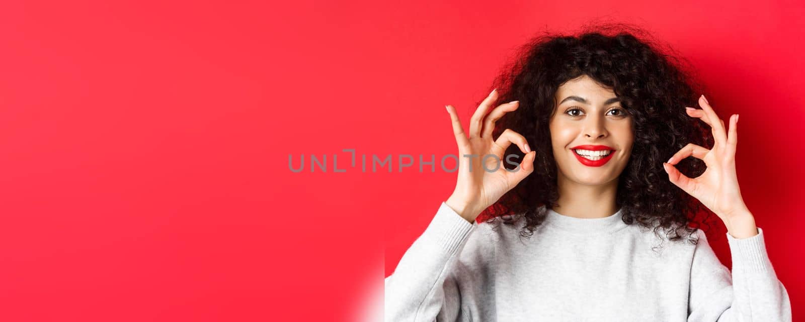 Close-up portrait of smiling woman with curly hair and red lips, showing okay gesture and looking satisfied, praise good product, standing on red background.