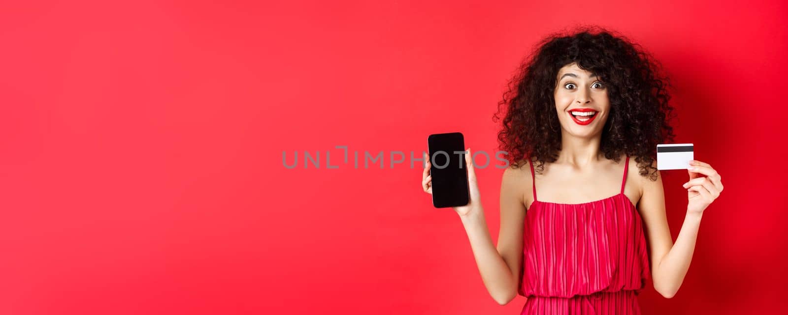 Excited lady in red dress showing plastic credit card and smartphone screen, smiling amazed, standing on studio background by Benzoix