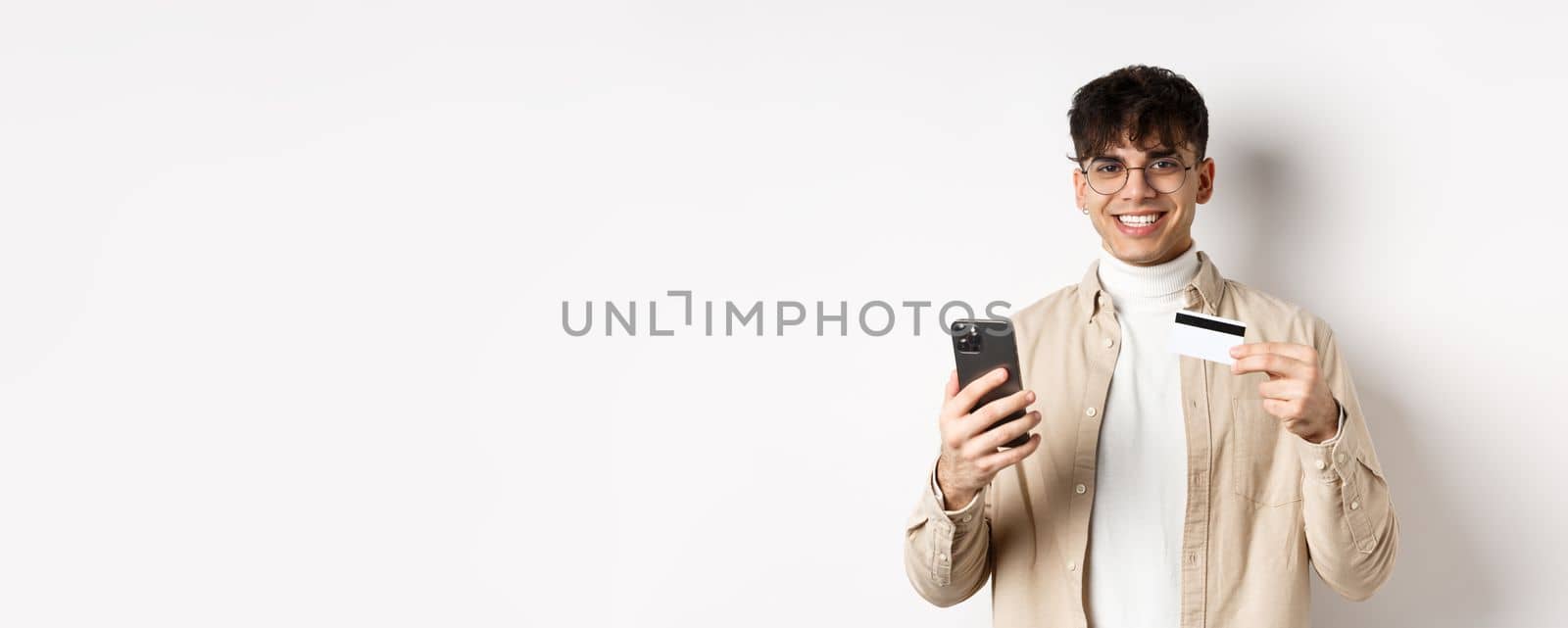 Portrait of natural young man in glasses paying in internet, showing smartphone and plastic credit card, standing on white background.