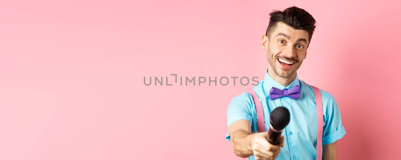 Smiling guy with moustache and bow-tie asking for comments or interview, stretch out hand with microphone, standing on pink background.