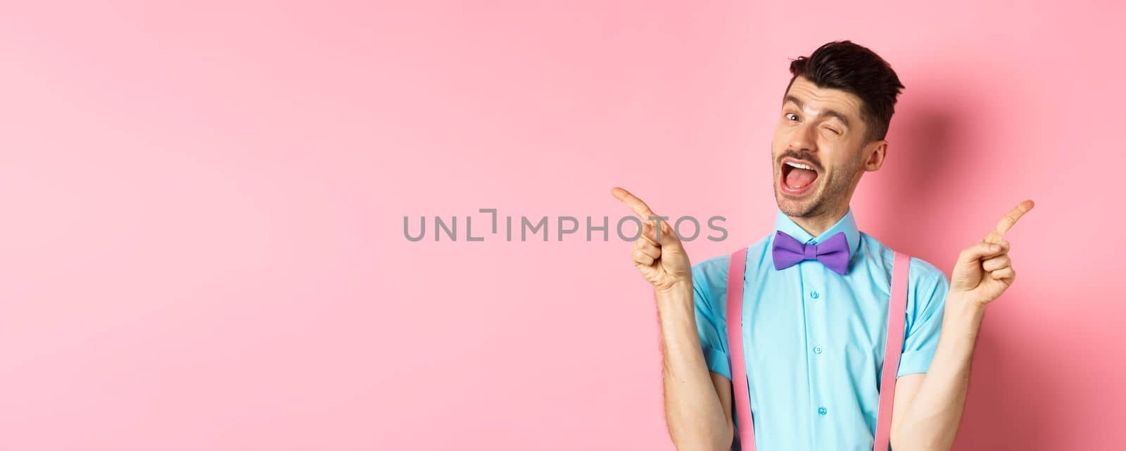 Cheerful young man smiling and winking, showing two sides, pointing fingers sideways and recommending promo offers on pink background.