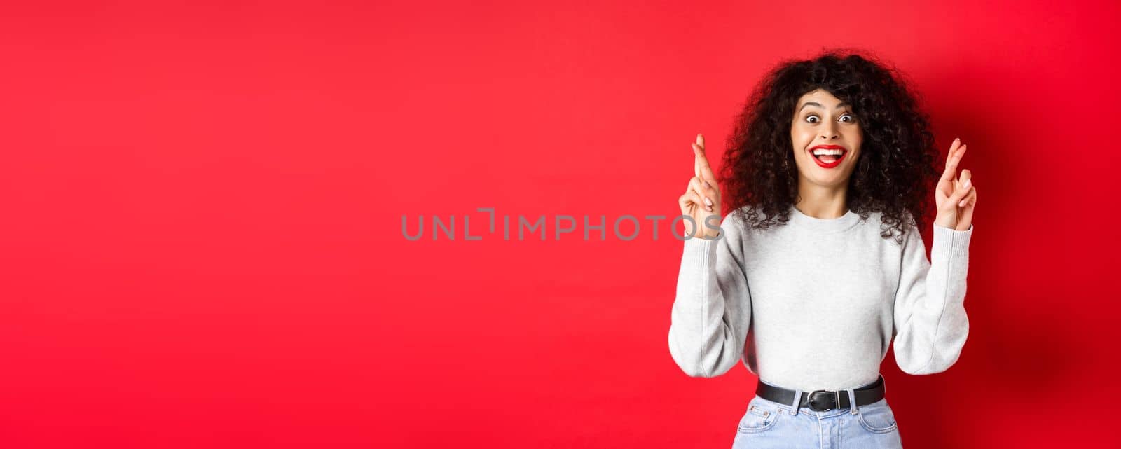 Hopeful excited woman making a wish, cross fingers for good luck and smiling amazed at camera, praying for dream come true, waiting for results, red background by Benzoix