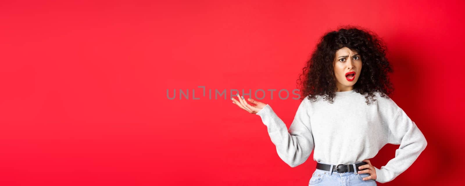 Annoyed and confused caucasian woman with curly hair, raising hand up and arguing, having argument, cant understand something, standing on red background by Benzoix