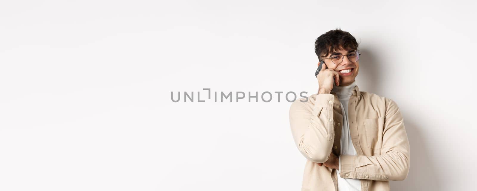 Real people concept. Happy young guy talking on mobile phone, look aside and holding smartphone near ear, standing on white background.