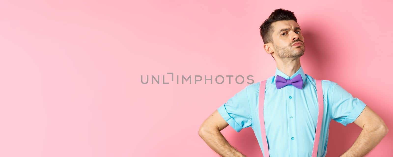 Proud and confident caucasian guy with moustache and bow-tie, looking arrogant with chin up, frowning and looking at camera, standing on pink background.