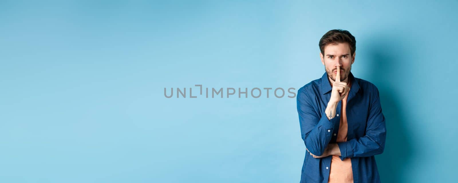 Angry man shushing, press finger to lips and frowning, tell to be quiet, show taboo gesture, standing on blue background.