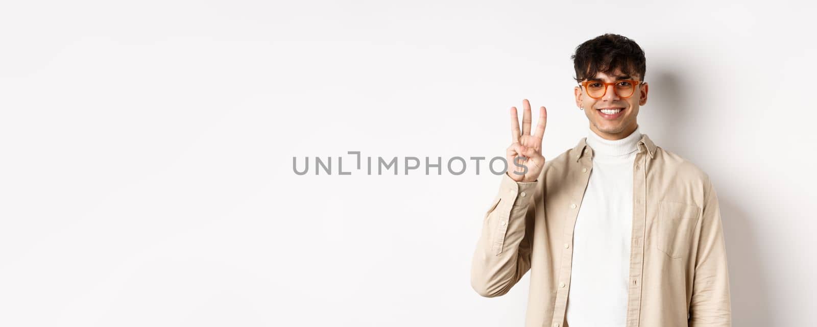 Handsome caucasian guy in glasses showing three fingers and smiling, standing on white background by Benzoix