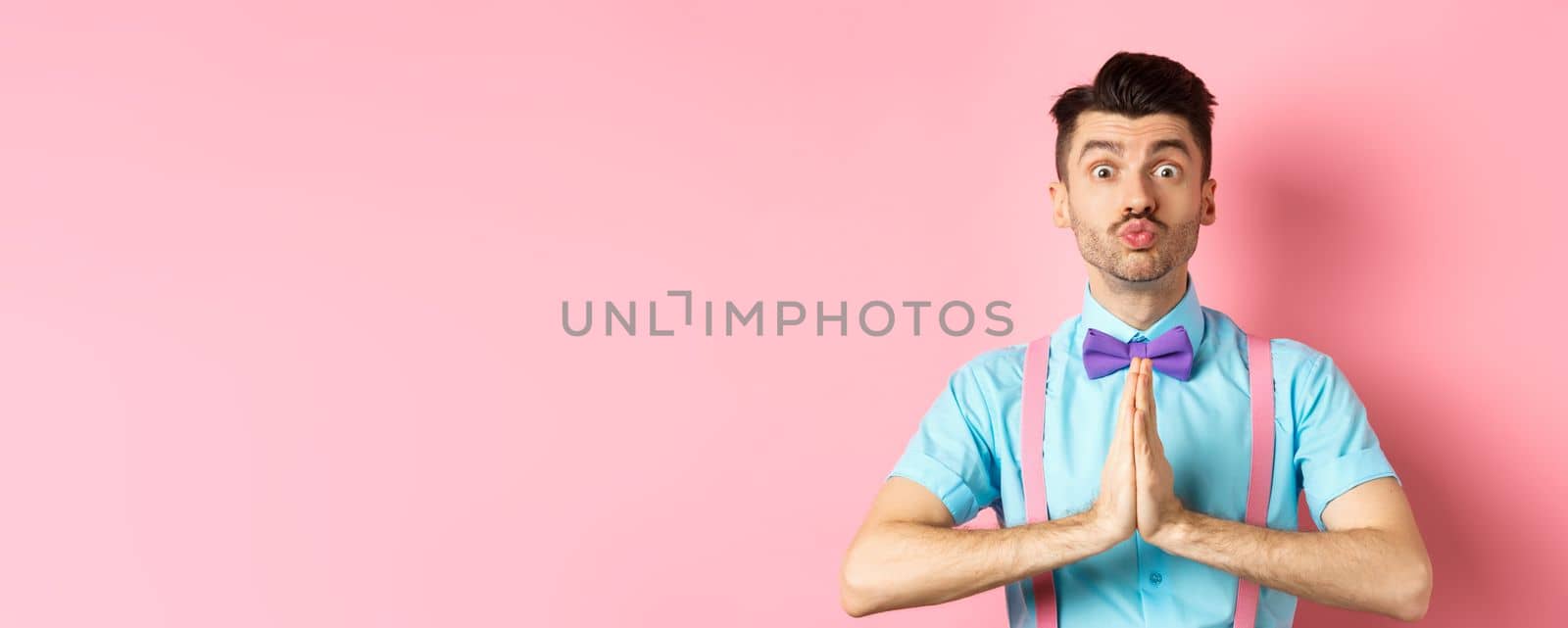 Young guy with cute face pucker lips and asking please, begging for favour, need something and lookin gpleading at camera, standing over pink background.