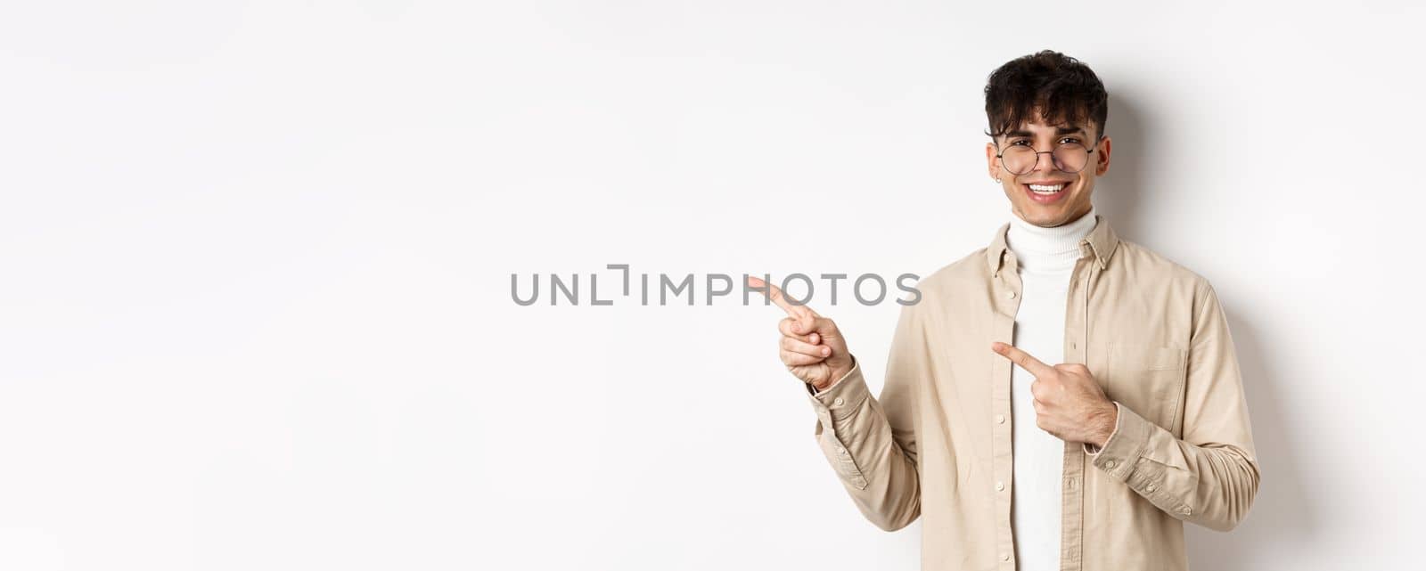 Portrait of handsome caucasian young man in glasses showing logo, smiling and pointing fingers left, inviting to check out promo deal, white background.