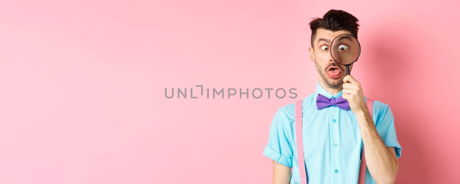 Funny man in bow-tie look through magnifying glass, squinting and making silly faces, standing on pink background by Benzoix