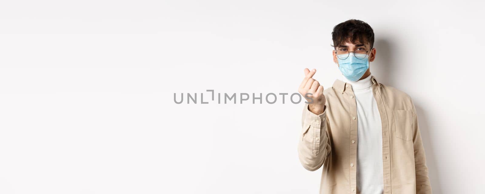 Covid-19, health and real people concept. Cute young man in medical mask showing finger heart and look at camera, standing on white background by Benzoix