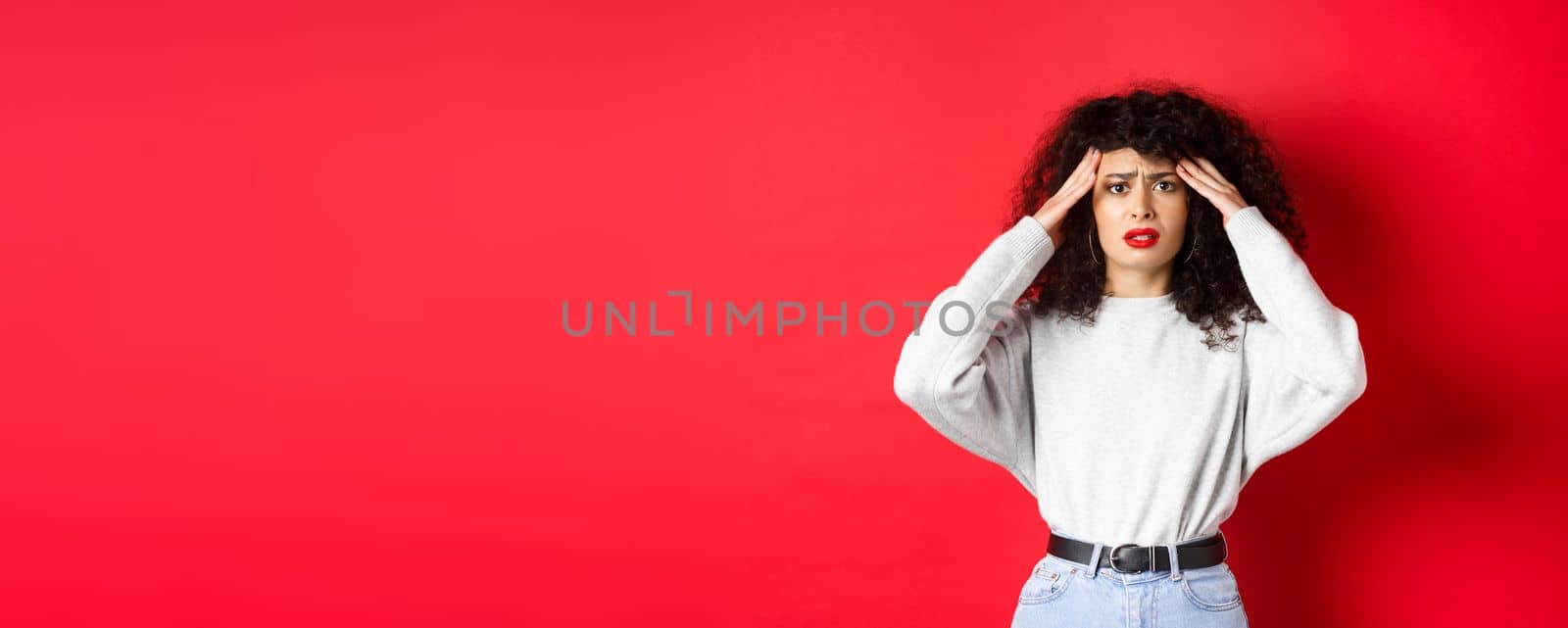 Worried and concerned young woman touching head and grimacing, having headache, standing with migraine on red background.