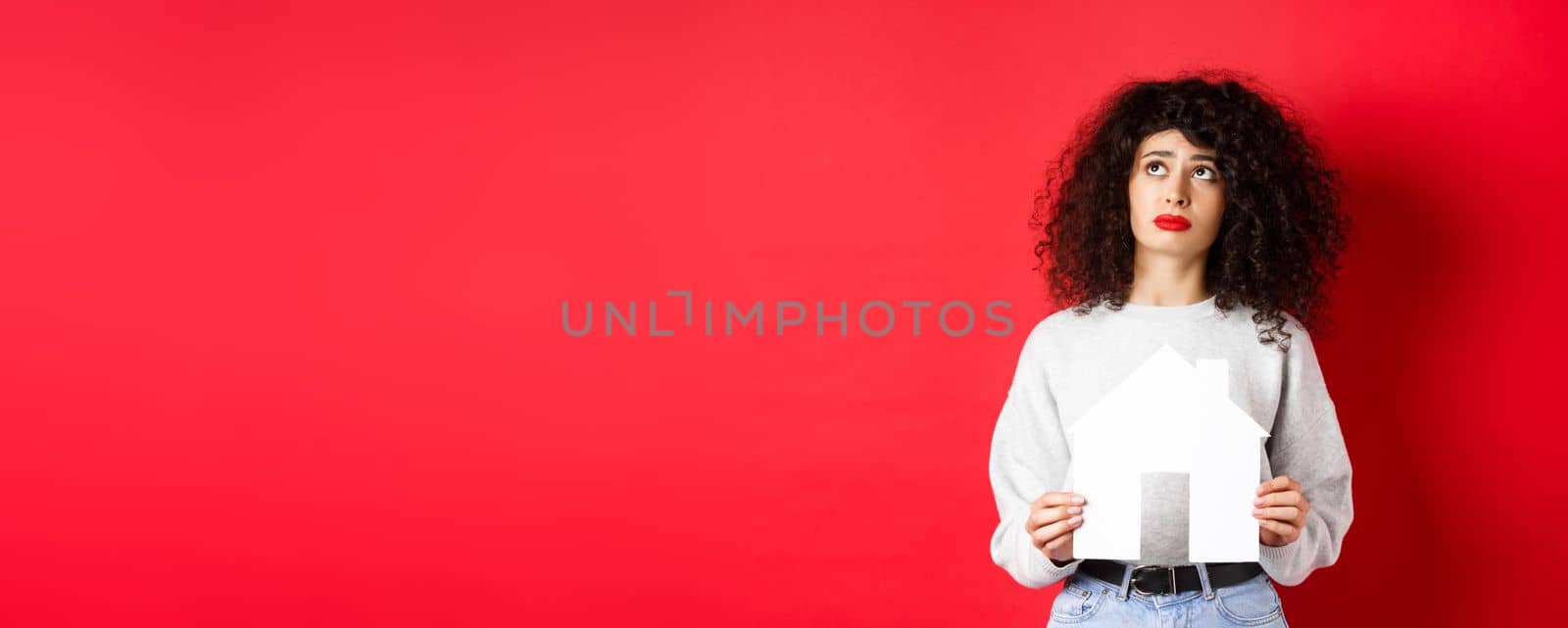 Real estate. Sad woman dreaming of buying apartment, holding paper house cutout and looking up distressed, standing on red background by Benzoix