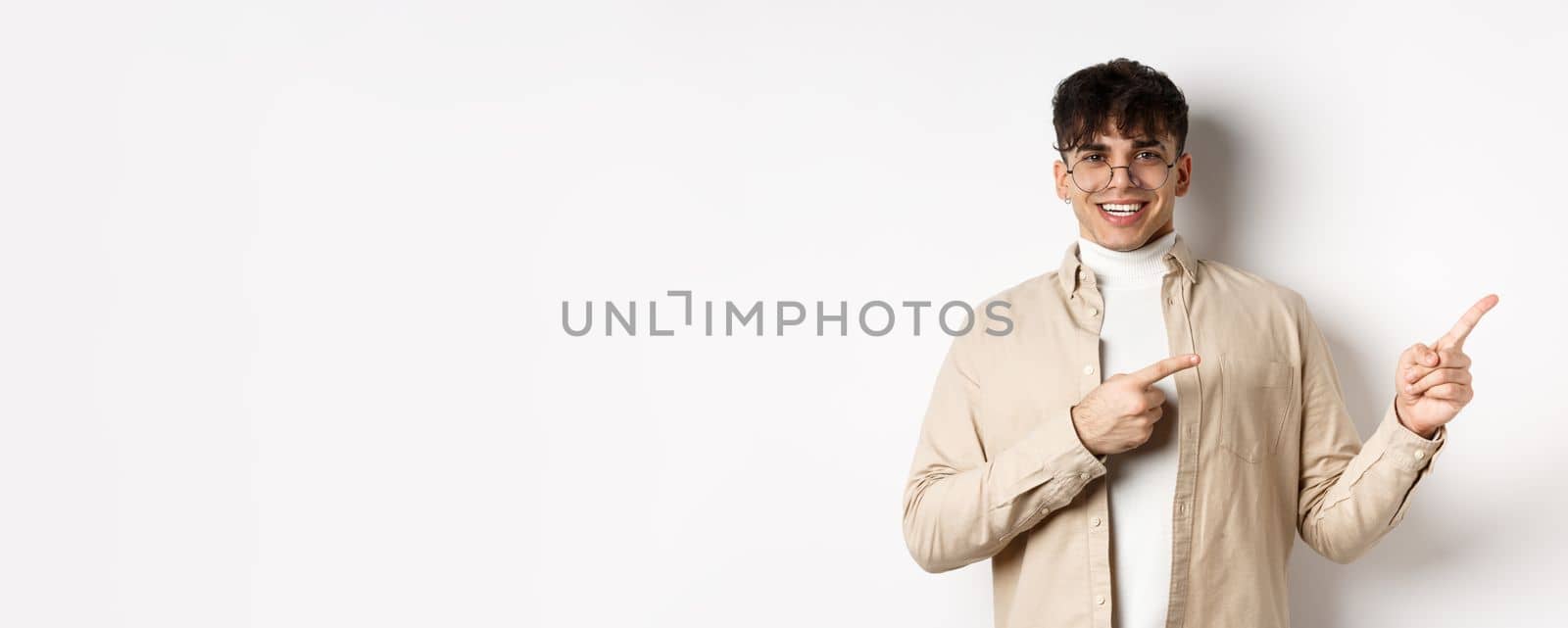 Proud and happy young man in glasses showing logo, pointing fingers at upper right corner and smiling, standing on white background.
