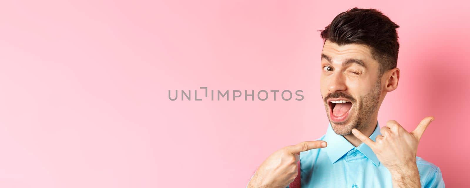 Cheeky and funny guy showing phone gesture, asking to call him and winking, telling dial his number, standing over pink background by Benzoix