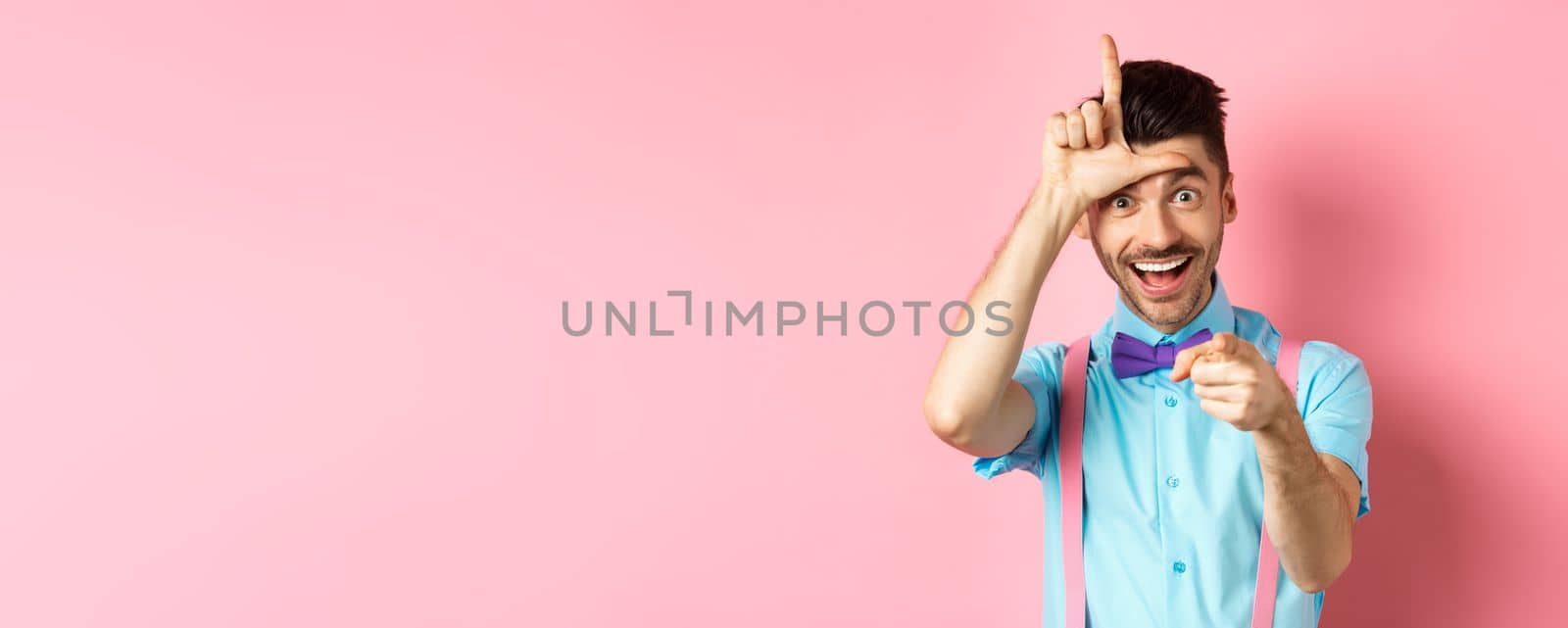 Image of funny guy pointing at you camera and showing loser sign, mocking person who lose, standing over pink background.