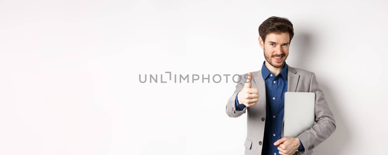Successful businessman in stylish suit carry laptop and looking confident at camera, showing thumbs up in approval, white background.