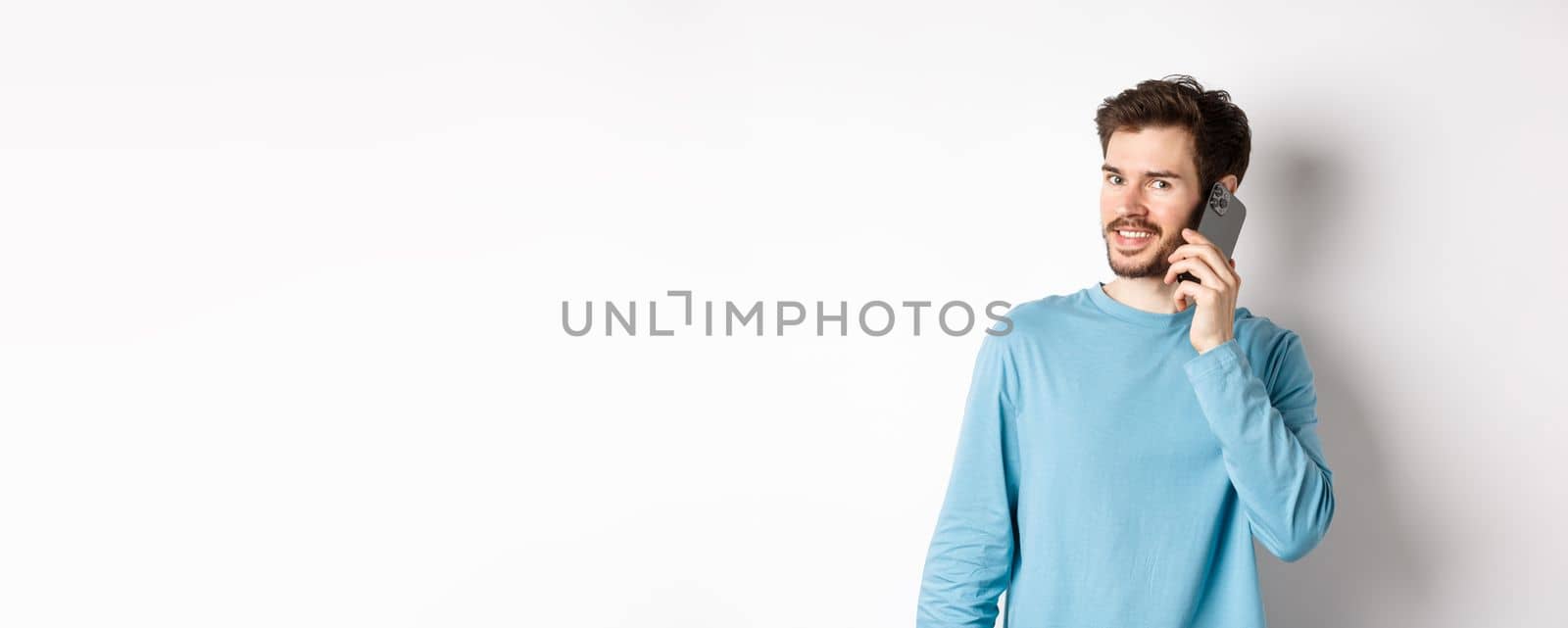 Technology concept. Young male model talking on mobile phone, calling someone on smartphone and smiling, standing over white background.