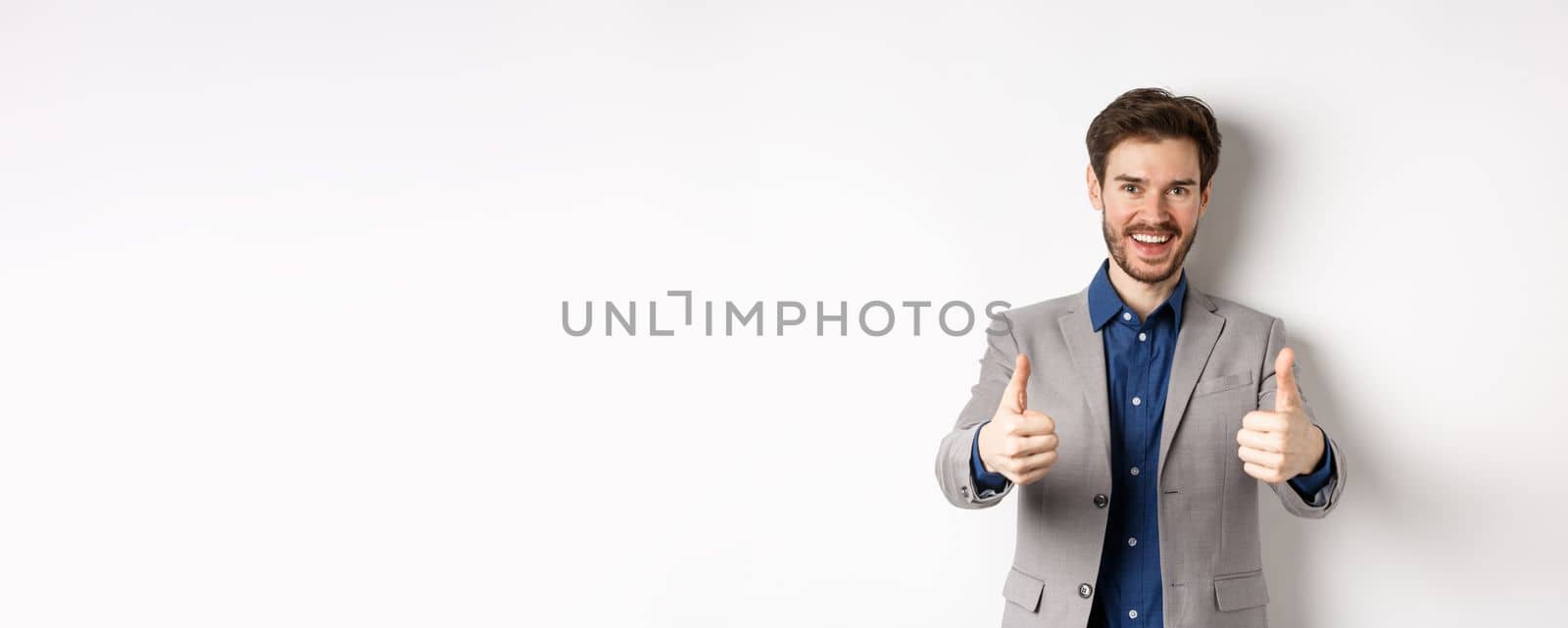 Cheerful and positive young man in business suit showing thumbs up and smiling, like something good, praise good job, standing happy on white background.