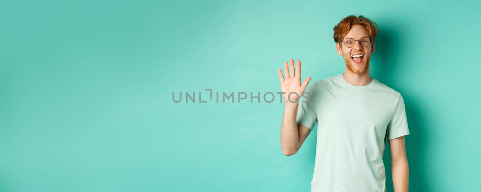 Friendly bearded guy in glasses saying hello, waving hand to greet and welcome you, standing cheerful and smiling over turquoise background by Benzoix