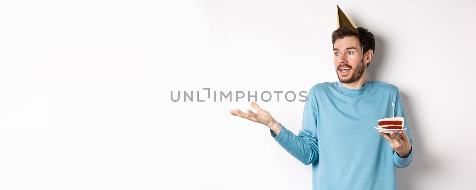 Celebration and holidays concept. Surprised man shrugging in disbelief, looking away, holding birthday cake and wearing party cone hat, white background by Benzoix