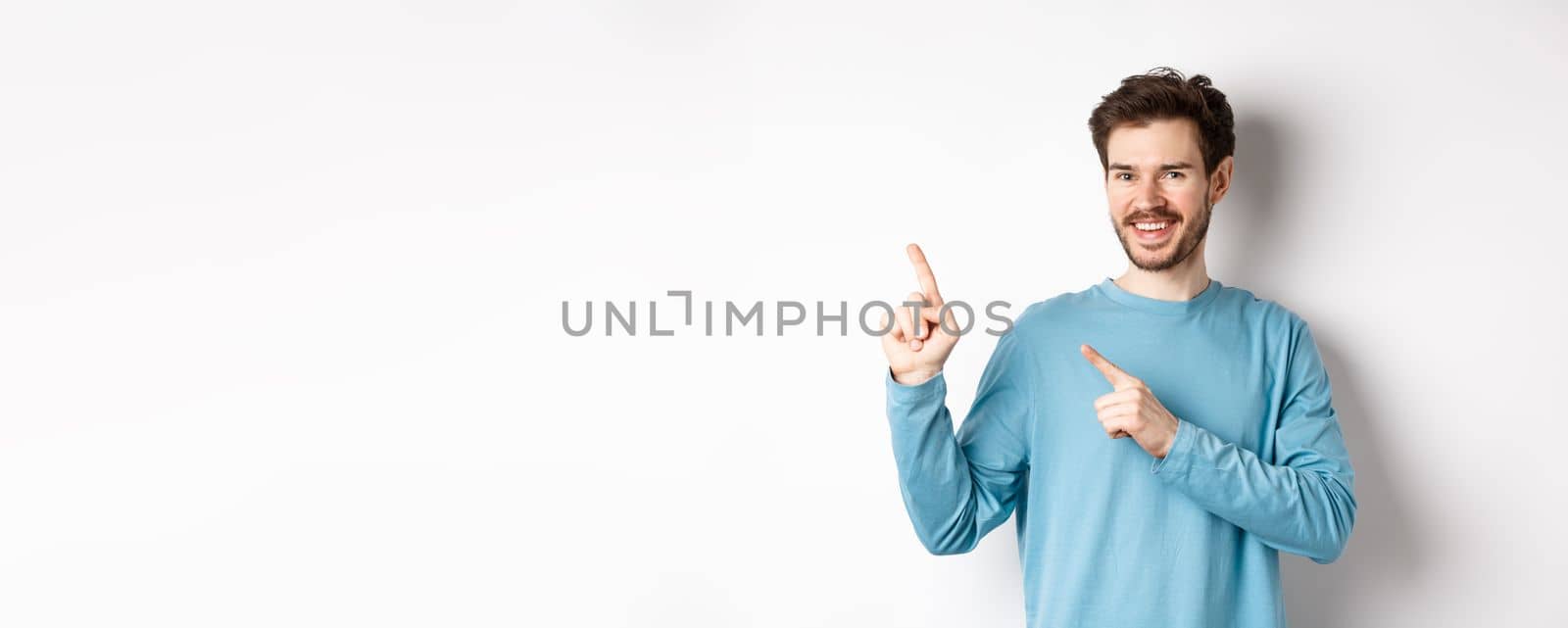 Cheerful bearded man showing advertisement on copy space, pointing fingers at upper left corner logo and smiling, standing over white background by Benzoix