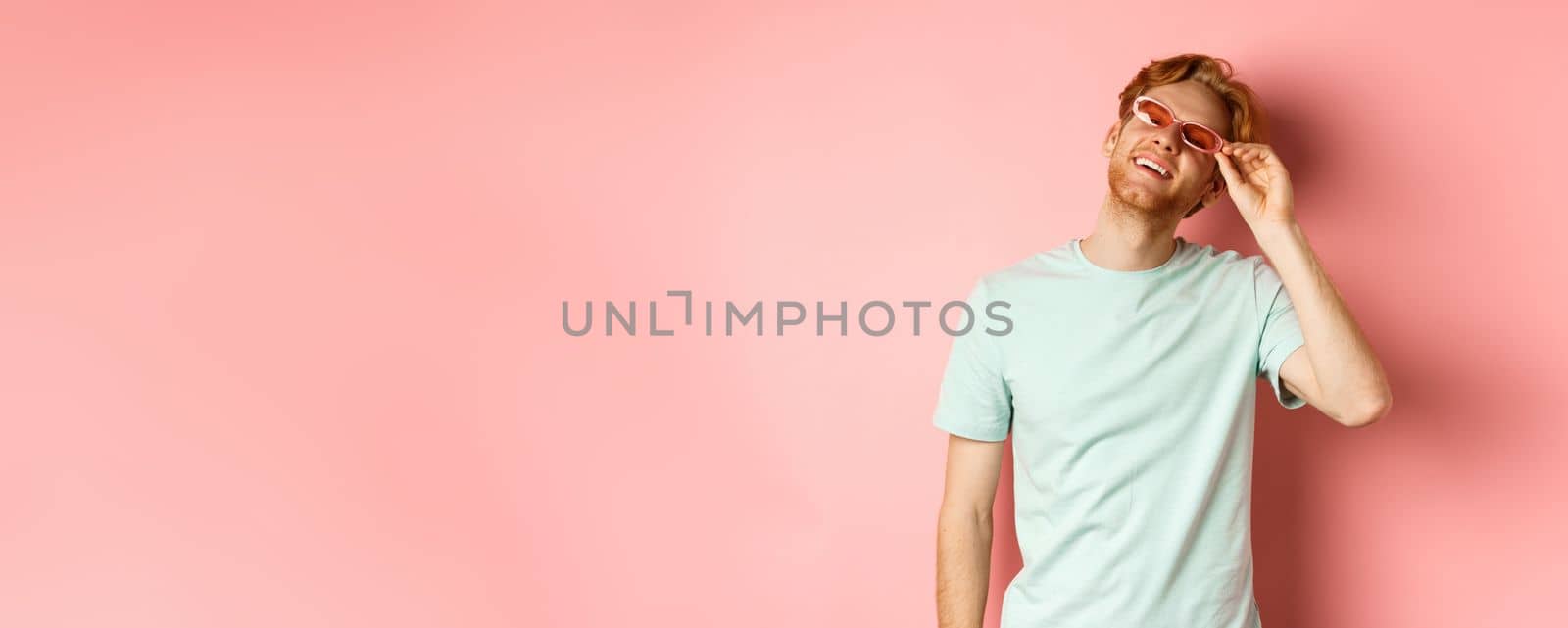 Tourism and vacation concept. Happy young man with red hair relaxing, wearing sunglasses and smiling carefree, standing over pink background.