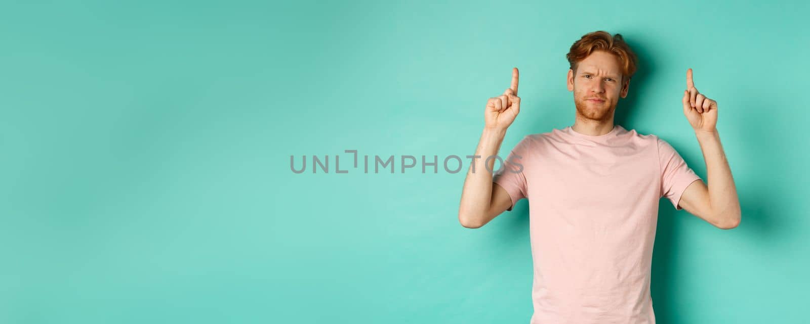 Skeptical and disappointed redhead man pointing fingers up, frowning displeased and looking upset, standing over turquoise background.